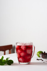 Cold refreshing hibiscus tea in a glass with ice, dry tea leaves of red herbal tea with lime in the background and fresh aromatic mint