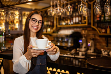 Portrait of a young waitress holding a cup of coffee in a cafe. Attractive waitress serving coffee. Maybe you want a cup of coffee Girl holding a drink stretching her palm hands forward