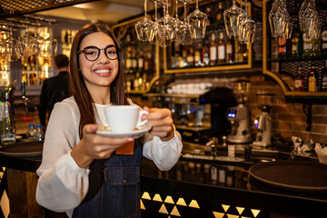Portrait of a young waitress holding a cup of coffee in a cafe. Attractive waitress serving coffee. Maybe you want a cup of coffee Girl holding a drink stretching her palm hands forward