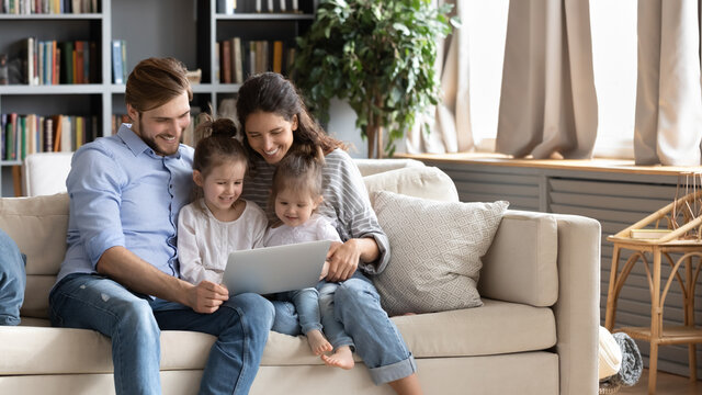 Wide banner panoramic view of happy young Caucasian family renters with two small daughters relax on sofa using laptop together. Smiling parents with little girls children browse internet on computer.