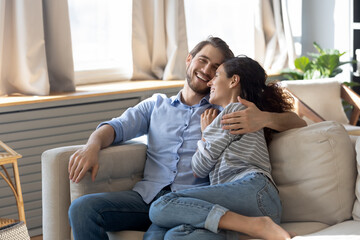 Fototapeta na wymiar Happy millennial Caucasian couple sit relax on comfortable couch at home hugging and cuddling. Smiling young man and woman rest on sofa in living room enjoy family romantic leisure weekend together.