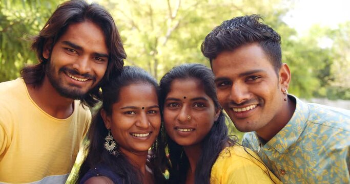 Four friends, two female sand two males, stand pose together under tree in nature park garden lawn for a camera selfie photo video pose with toothy smile and say cheese laugh