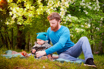 Naklejka na ściany i meble Father with child having picnic outdoors near apple tree with white flowers. Dad with one son relax in spring garden. Father and little boy in green park.