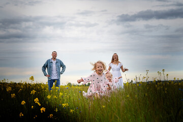 A family including two young parents and daughters on a walk in a meadow with grass and flowers. Dad, mom, girls relaxing and having fun in nature on a summer day with clouds