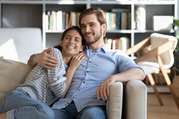 Happy young Caucasian couple sit rest on sofa hugging and cuddling, look in distance dream of future together. Smiling millennial man and woman relax on couch embracing, enjoying weekend.