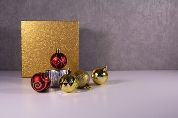 Gold and red christmas balls with golden gift box in front view style. Gray textured background. There is a free space for your design.