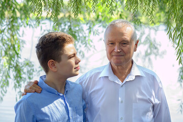 Portrait of happy сaucasian teen boy and elderly grandfather  in blue shirts and jeans sitting by the lake. Relaxing outdoors together. The concept of trust, happiness.  Older generation.