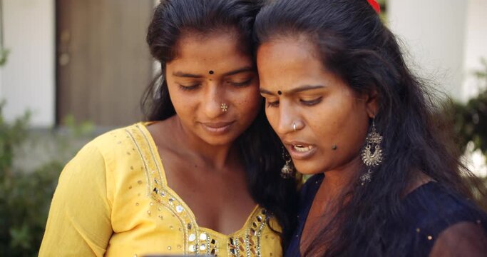 Two Indian women in traditional clothes in conversation teamwork as they look intently at a mobile phone screen and smile with joyous happiness slow-motion static shot 