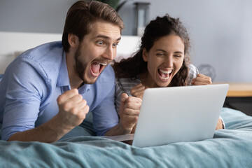 Overjoyed millennial man and woman lying in bed scream celebrate online victory or win on laptop. Excited young Caucasian couple feel euphoric triumph with good news on computer. Success concept.