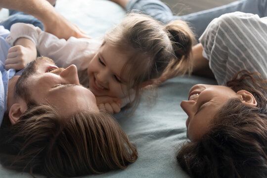 Close up of overjoyed young Caucasian parents lying in bed have fun play with little daughter. Smiling family with small girl child feel playful in bedroom, enjoy leisure weekend in own home or house.