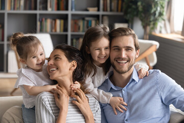 Happy small girls children hug excited young mom and dad show love and care relaxing on sofa at home together. Smiling little daughters embrace Caucasian parents, play in cozy living room on weekend.