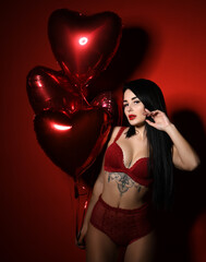 Beautiful sexy young woman with red heart balloons for birthday or valentines day party posing in underwear on dark red background 