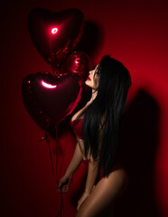 Beautiful sexy young woman with red heart balloons for birthday or valentines day party posing in underwear on dark red background 