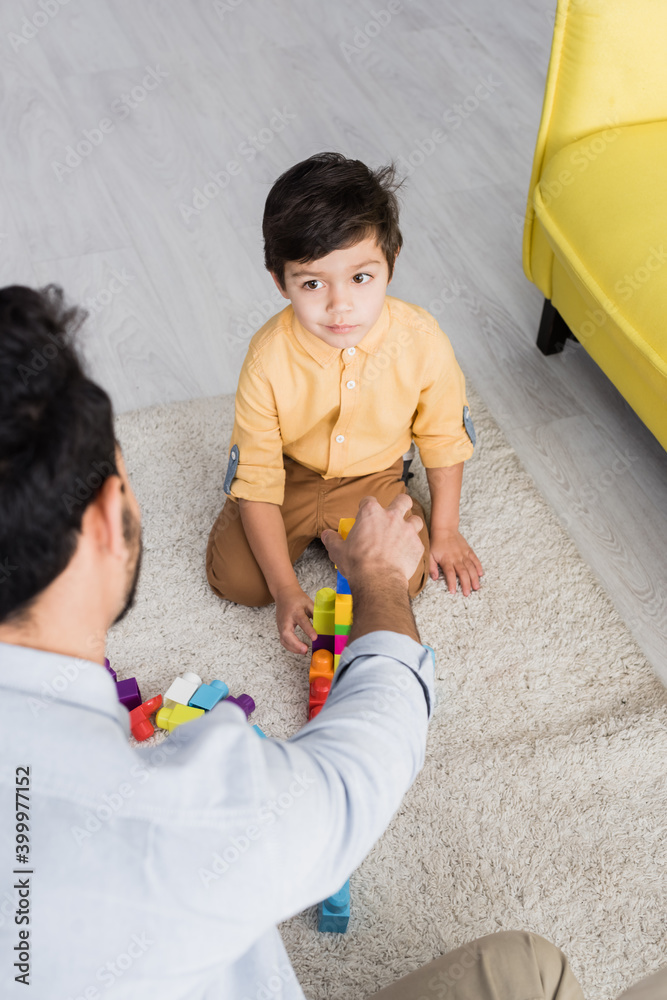 Wall mural Boy looking at father playing building blocks on blurred foreground at home, two generations of men