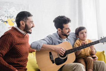 Smiling hispanic father playing acoustic guitar near son and grandfather at home, three generations of men