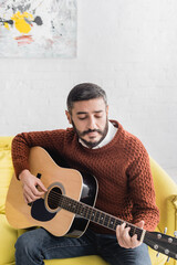 Hispanic man playing acoustic guitar while sitting on sofa at home