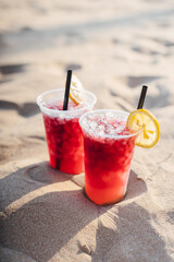 Tropical red ice cold drinks. A glass of cold drink stands on the sand. Drinking straw and orange slice. Hot summer and relaxation.