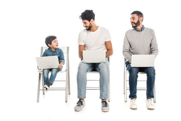 hispanic dad and grandfather looking at cheerful boy while using laptops together on white, three...