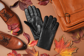 Stylish black leather gloves, pair of shoes, bag and dry leaves on brown background, flat lay