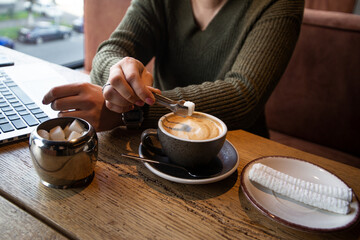 Woman in green pullover adds a piece of sugar to her cappucino with tongs while working at a coffee shop. Cozy cafe concept. Cut view of laptop on background.