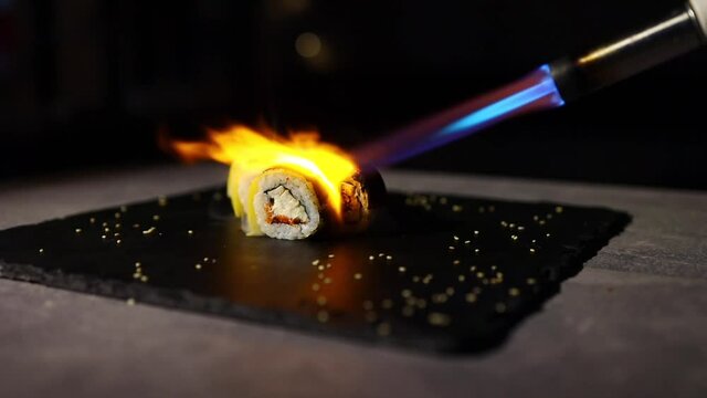 Sushi lies on a black sheet of iron and the cook bakes them with a gas burner using the aburi method. Close-up