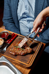 A waiter in a restaurant puts a steak for a customer using tongs on a wooden tray. Close-up of hands