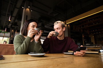 Young couple enjoys delicious coffee in cafe. They holding cappuccino cups and looking at each other. Tasty drinks. Coffee shop or restaurant on background.