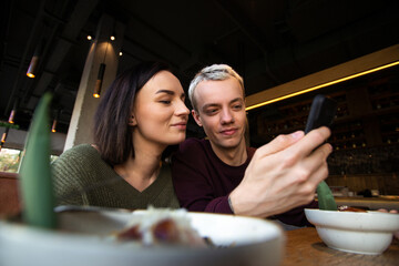 Man sharing good photos of food to woman. Blurred tasty meal on front. Stylish blonde man showing interesting message in his smartphone. Young couple spending time at cafe or restaurant.