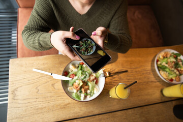 Woman takes photo of meal. Healthy food concept. Top view. Social networks concept. Caesar salad on white plate. Share her meal on social media account. Restaurant or cafe on background.