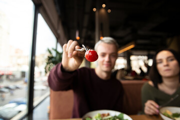 Focus on red tomato. Yummy salad. Blurred caucasian blonde man and brunette woman. Cafe of restaurant on background. Healthy lifestyle. More vegetables concept.