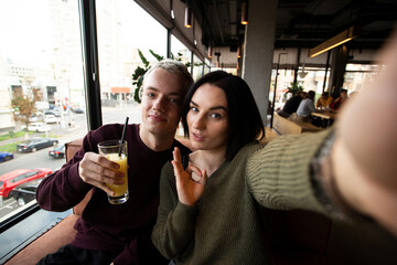 Young couple posing for selfie in public place. Attractive brunette woman and good looking blond man take photo in a restaurant together. Man holds a glass of orange juice. Woman makes okay gesture.