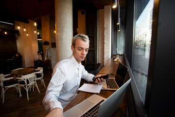 Fototapeta na wymiar Freelance programmer sits near the window in a coffee shop and works. Stylish blonde man in white shirt uses laptops. Cozy coffee shop atmosphere. Cafe or restaurant on background.