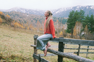 Fototapeta na wymiar Portrait of woman wearing dreadlocks sitting on wooden fence in autumn mountain village forest