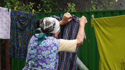 Great-grandmother with traditional head scarf on her head spreading and hanging colorful washed wet...