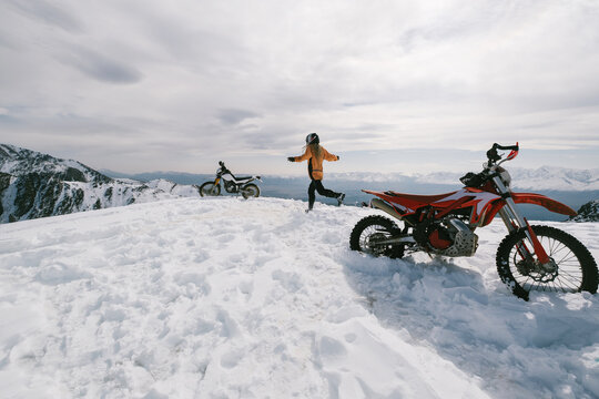 Female motorcyclist walking near her enduro motorcycle on snowy mountain top, snow peaks skyline view, high beautiful mountains