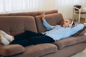 Rest at home after work during quarantine. Mature man with gray hair lies on the sofa and listens to music in his smartphone. Leisure time. Modern technology concept.