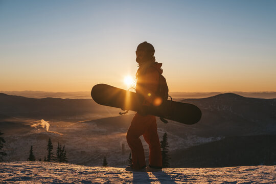 Female with snowboard. Snowboarder standing and resting in winter forest watching beautiful sunset and mountains landscape