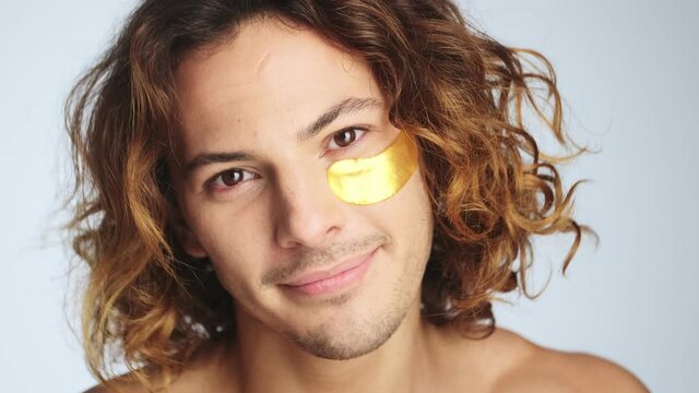 A handsome man is putting off hydrogel eye-patches while looking to the camera standing isolated over white wall in studio