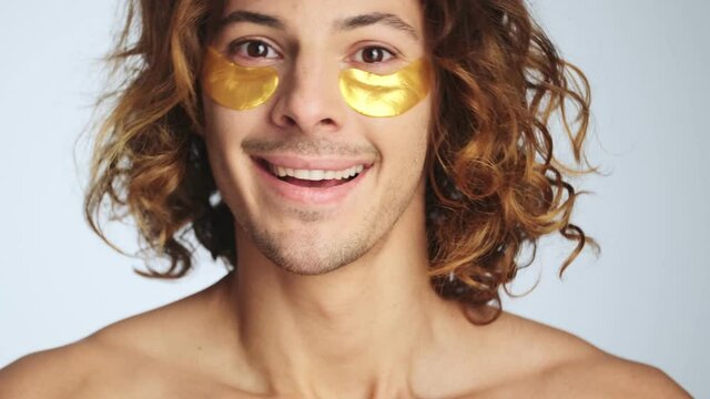 A pleased man with hydrogel eye-patches standing isolated over white wall in studio