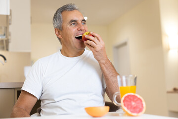 Happy adult man eating fruits and healthy food on breakfast. Morning routine concept. Cut halves of grapefruit and glass of orange juice on table. Cozy modern kitchen on background.