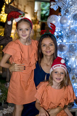 Single mother and two daughters by the Christmas tree wearing festive hats. Merry Christmas and happy holidays