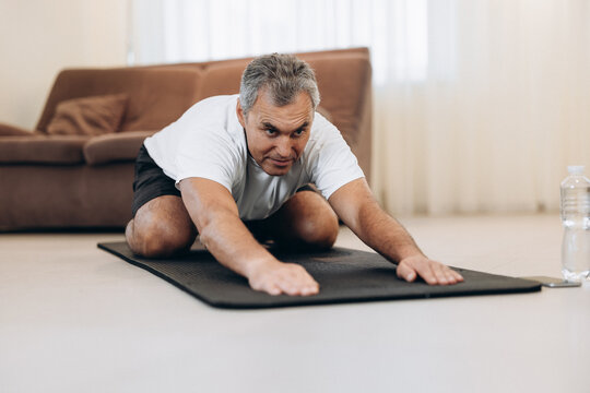 Old Man In Sportswear Lying On Mats Doing Child Asana, Exercise Calms Body And Mind, Stretching Hands Forward. Practicing Yoga At Home. Wellness Concept. Man Training To Have Flexible Body.