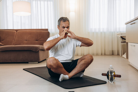 Elderly Sick Man With Runny Nose Sitting On Black Yoga Mat. Stay Home Concept. Man Blows His Nose After Workout Or Fitness. Practicing Sports At Home If You Feel Sick. Workout With Sports Equipment.