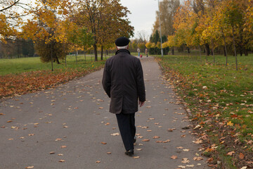 An old lonely pensioner man in a cap walks through the autumn park. View from the back.