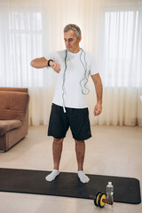 Fit man monitors his heart rate with a smart watch. Workout at home concept. Man with blue jumping rope around neck, standing on yoga mat and looking at his watch. Morning fitness in living room.