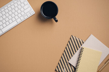 Modern office desk with keyboard, cup of coffee, paper notebooks and notepad on pastel brown...