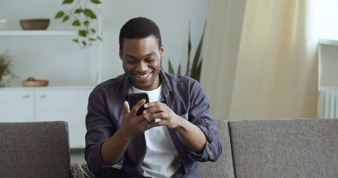 Mixed race guy afro american black man sitting at home in living room on couch learns good news happy to win lottery feels triumph watching tv calls on phone share emotions speaks on smartphone gadget