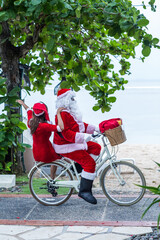 Fototapeta na wymiar Santa Claus and his wife Mrs. Claus wearing sunglasses and riding a bicycle in hot tropics by the ocean. Alternative exotic Christmas and New Year celebration