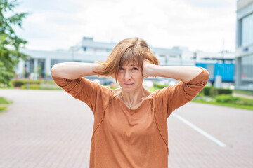 A middle-aged redhead woman in casual clothes stands on the street covering her ears with her...