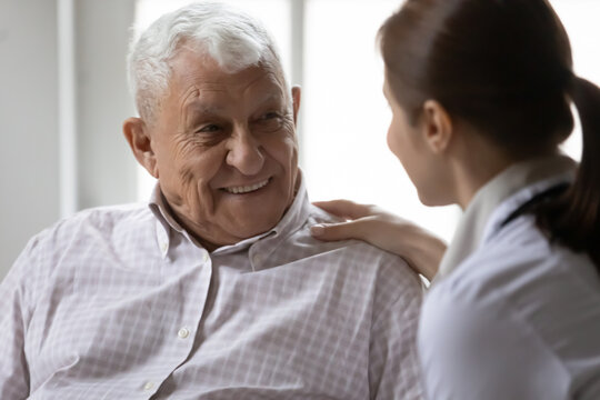 Young woman attending physician caring elderly aged clinic patient with sympathy talk ask about complaints. Pleasant female doctor gp hold hand on shoulder of smiling old man give hope tell good news
